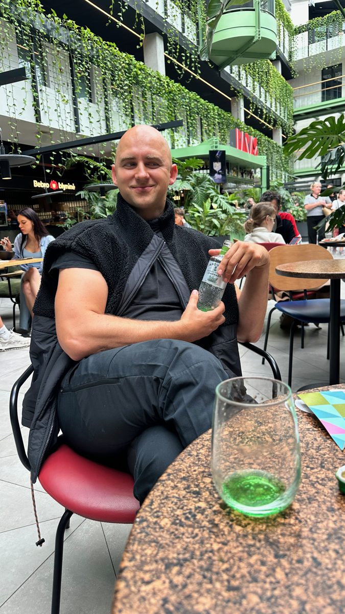 Lee at a cafe table under hanging plants, a green bottle in front of him.